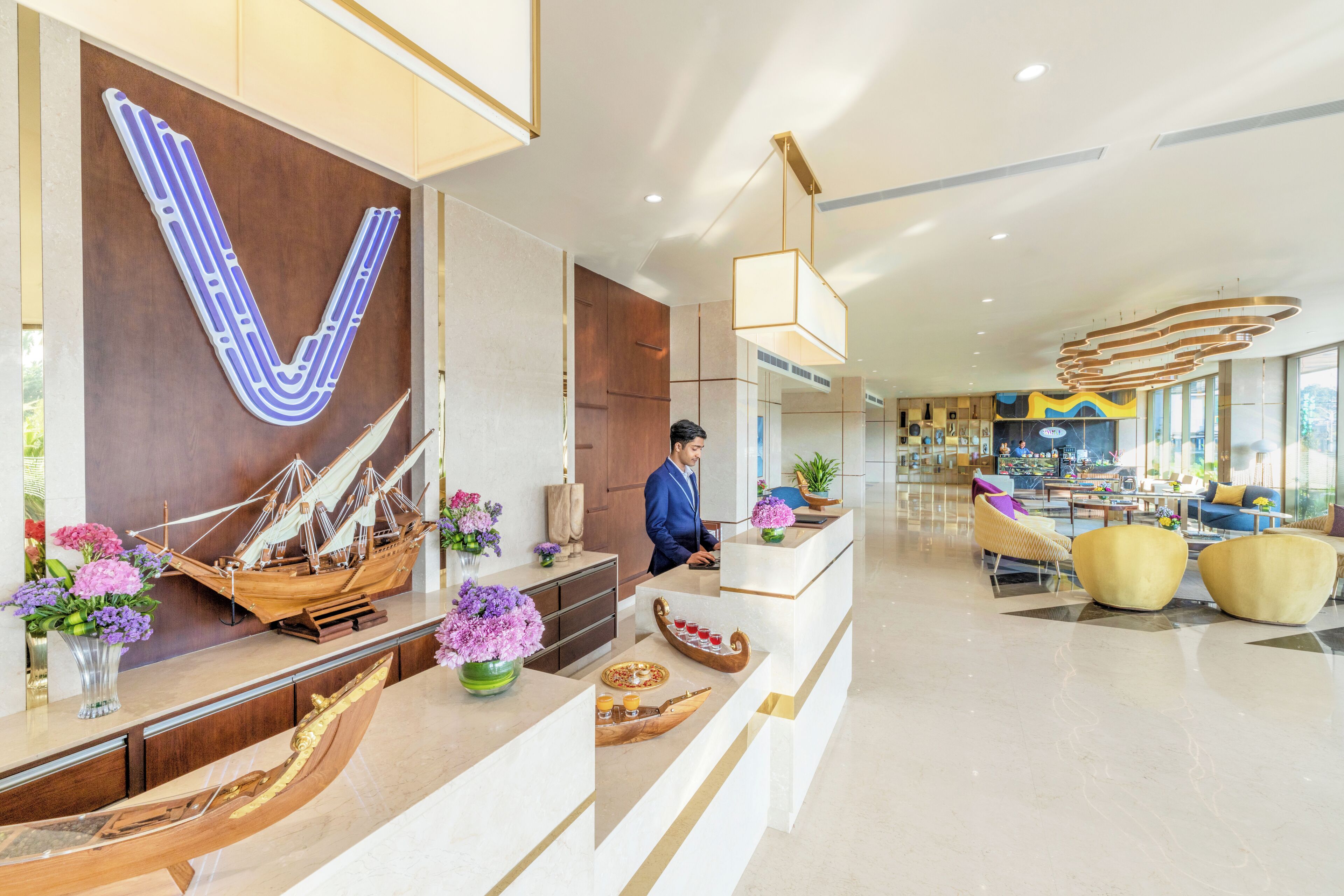 Un moderno vestíbulo de hotel con una recepción, una gran letra decorativa 'V' y una zona de estar con sillas amarillas.