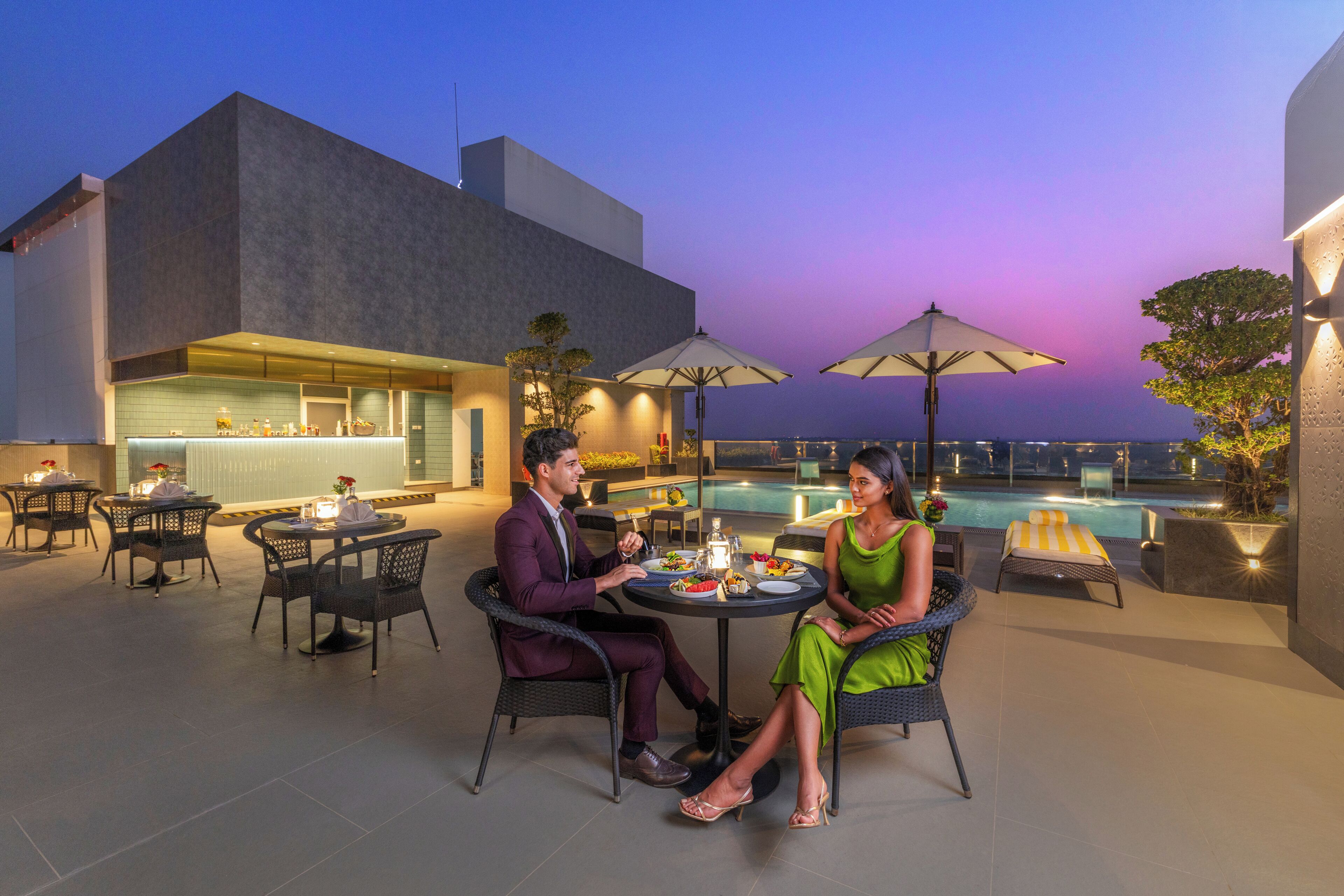 Una pareja cenando en una mesa al aire libre con vista a una piscina y un edificio moderno.