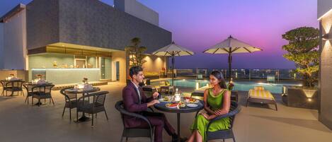Una pareja cenando en una mesa al aire libre con vista a una piscina y un edificio moderno.