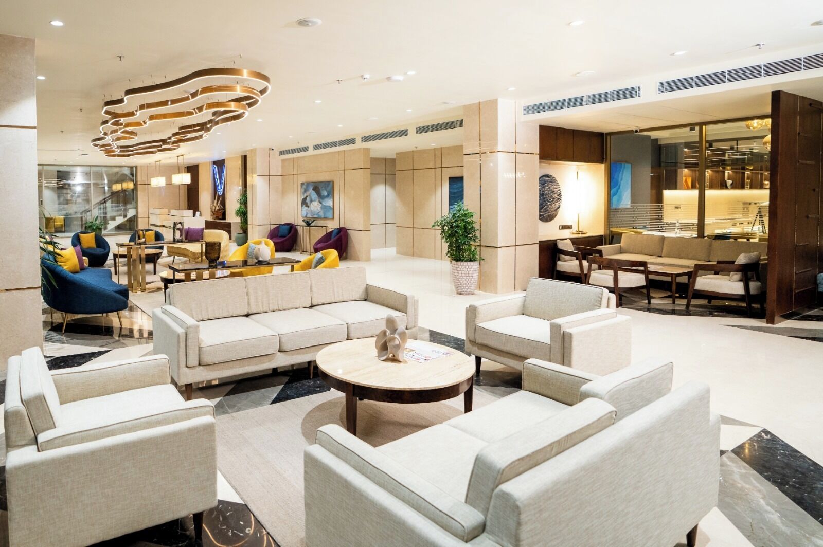 A modern hotel lobby with a sectional sofa, armchairs, a coffee table, and a dining area with a chandelier.