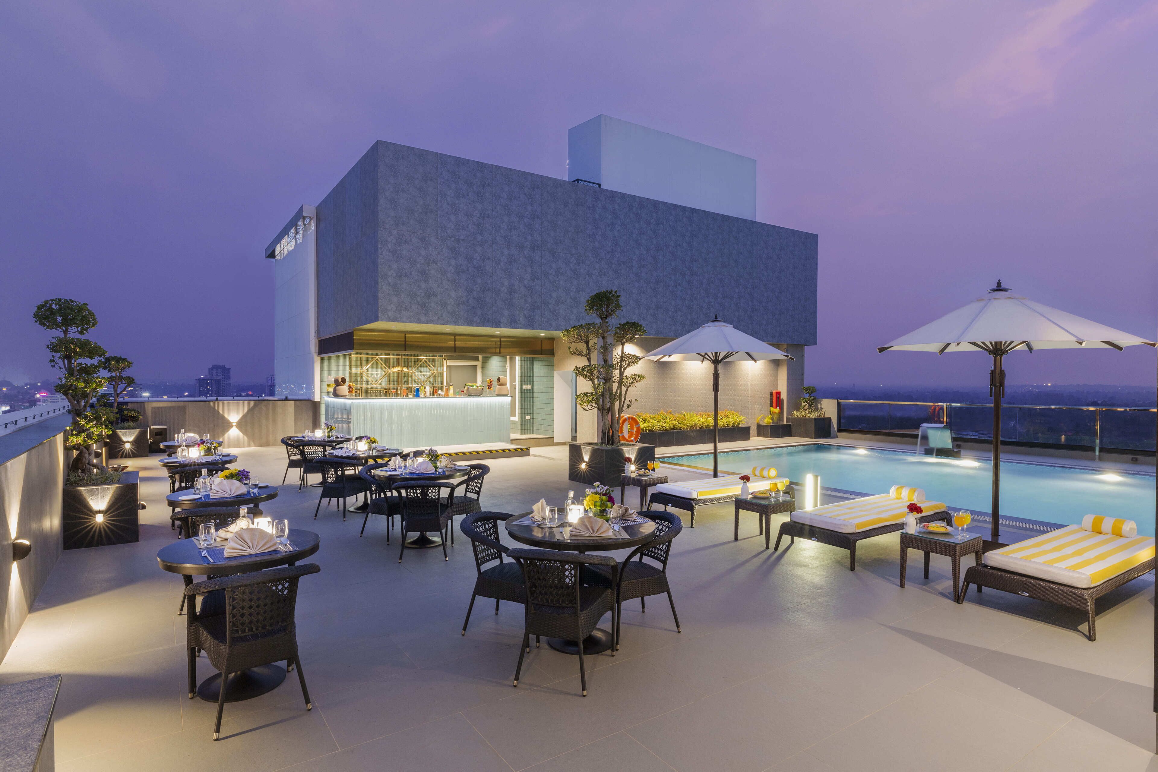 Un bar en la azotea con mesas y sillas, una piscina y un edificio moderno al fondo.