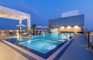 Ett poolområde på taket med en stor pool, parasoller, solstolar och en modern byggnad i bakgrunden.