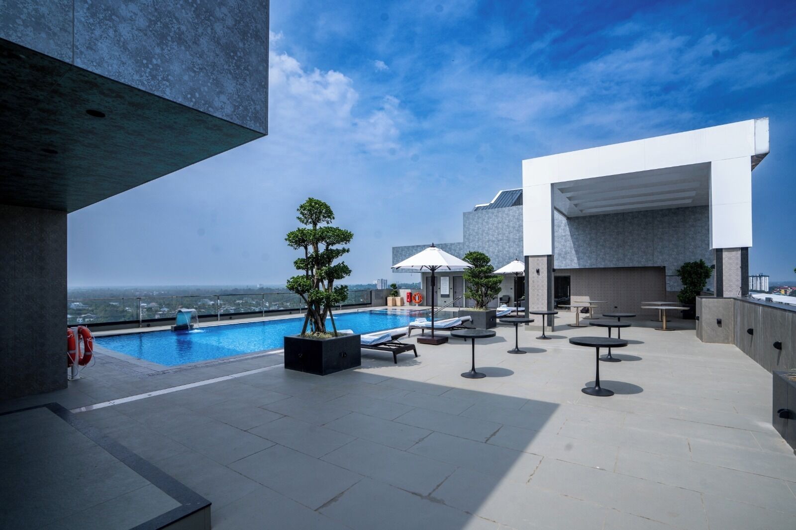 A rooftop pool area with a clear view of the city skyline, lounge chairs, umbrellas, and a small tree planter.