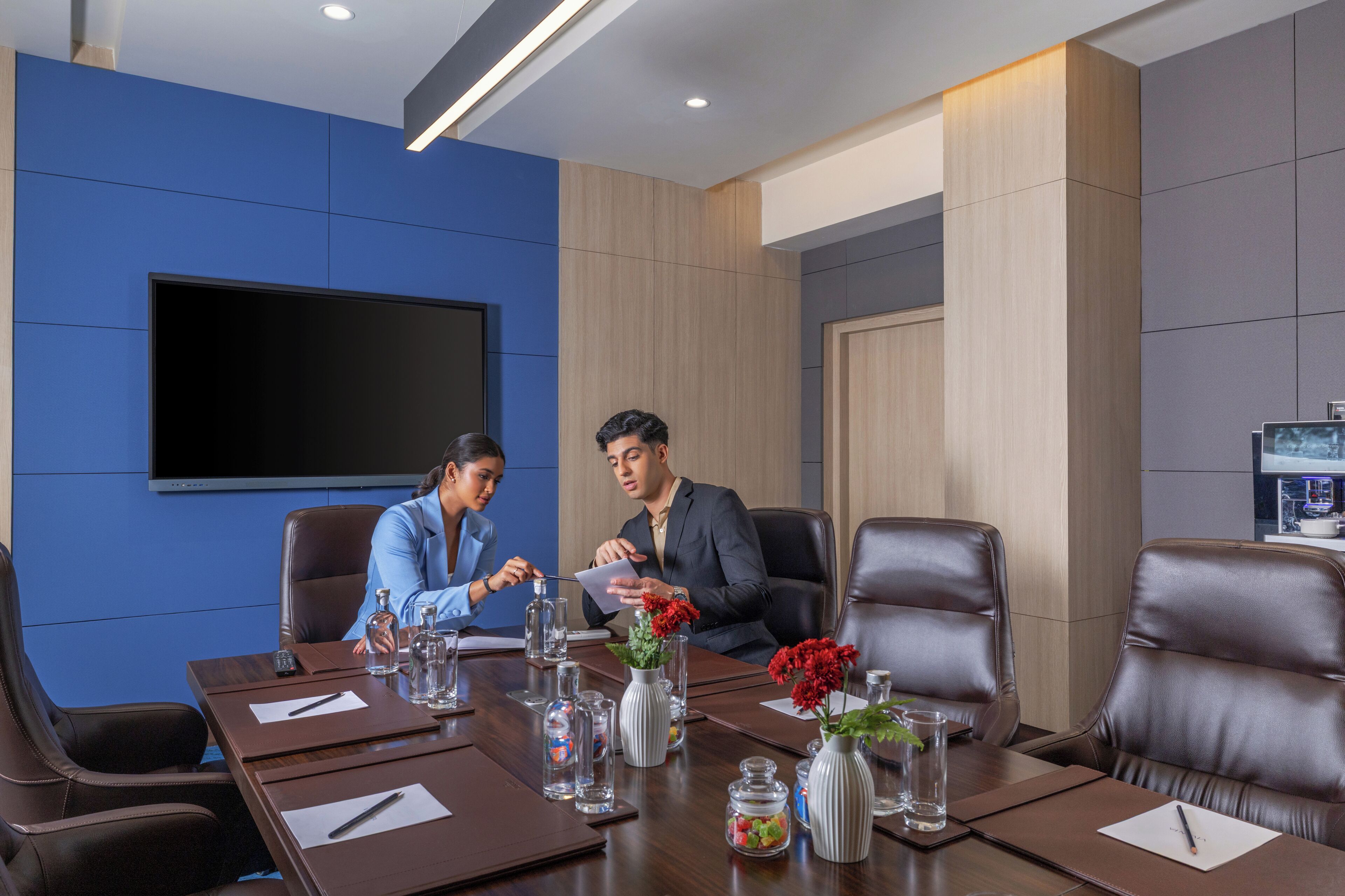 A conference room with a large flat-screen TV, leather chairs, a wooden table with water bottles, and vases with flowers.