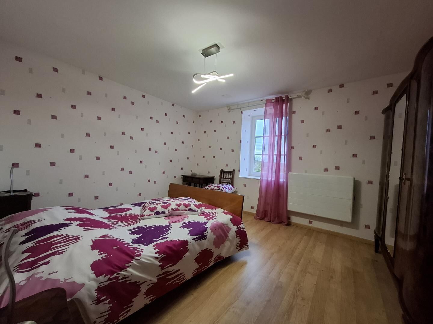 A bedroom with a bed, a radiator, a window with curtains, and a ceiling light fixture.