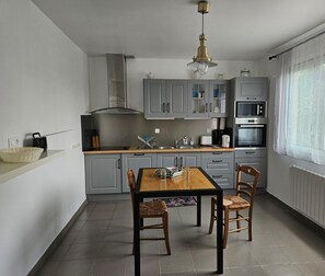 Eine moderne Küche mit grauen Schränken, einem Holzes Esstisch und einem Fenster mit durchsichtigen Vorhängen.