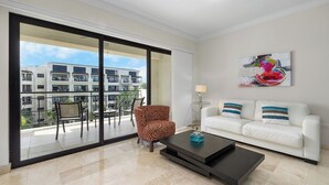 Una sala de estar moderna con un sofá blanco, un sillón estampado, una mesa de centro y un ventanal con vistas a un edificio y vegetación.