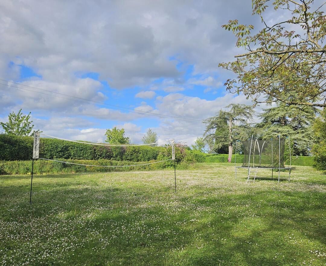 Un court de tennis avec un filet, entouré d’une zone herbeuse agrémentée d’arbres et d’une haie en arrière-plan.