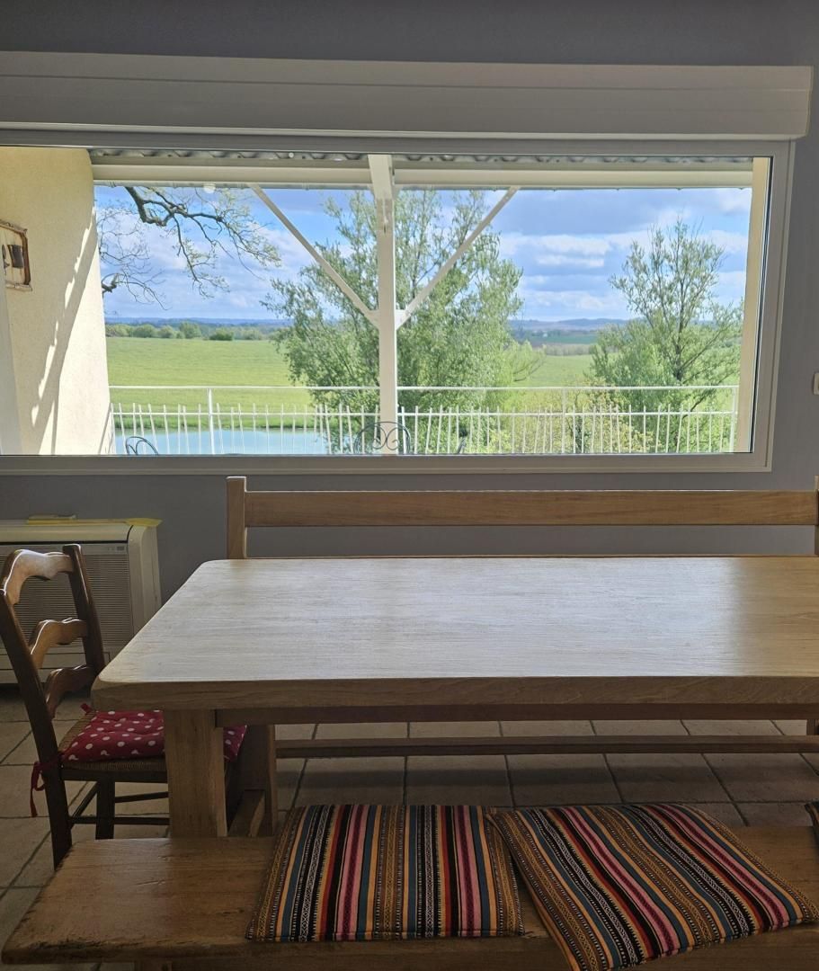 Un coin repas avec une grande table et des chaises, une vue sur un paysage verdoyant par la fenêtre et un petit tapis sur le sol.