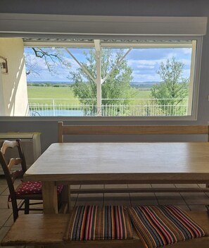 Un coin repas avec une grande table et des chaises, une vue sur un paysage verdoyant par la fenêtre et un petit tapis sur le sol.