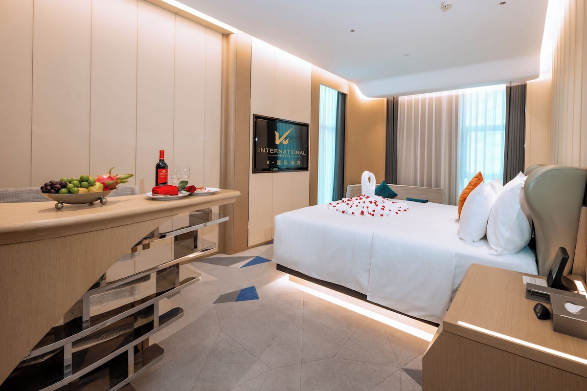A modern hotel room with a bed, bedside table, and a desk with a fruit bowl and wine glasses.