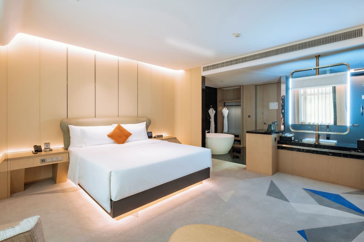 A modern hotel room with a large bed, bedside table, and a bathroom with a bathtub and mirror.