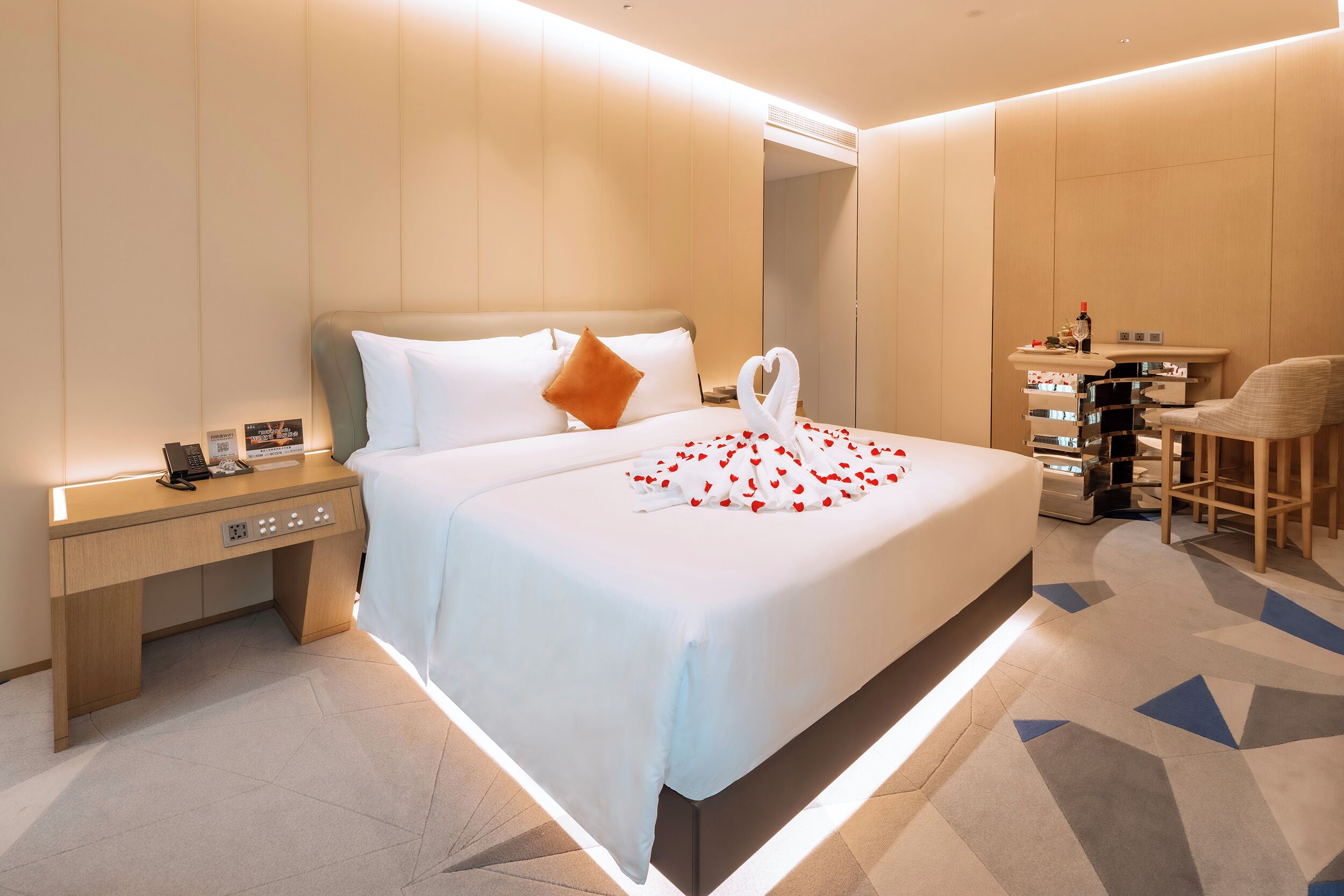 A modern hotel room with a large bed, a bedside table with a phone, a desk with a chair, and a small table with a mirror and toiletries.