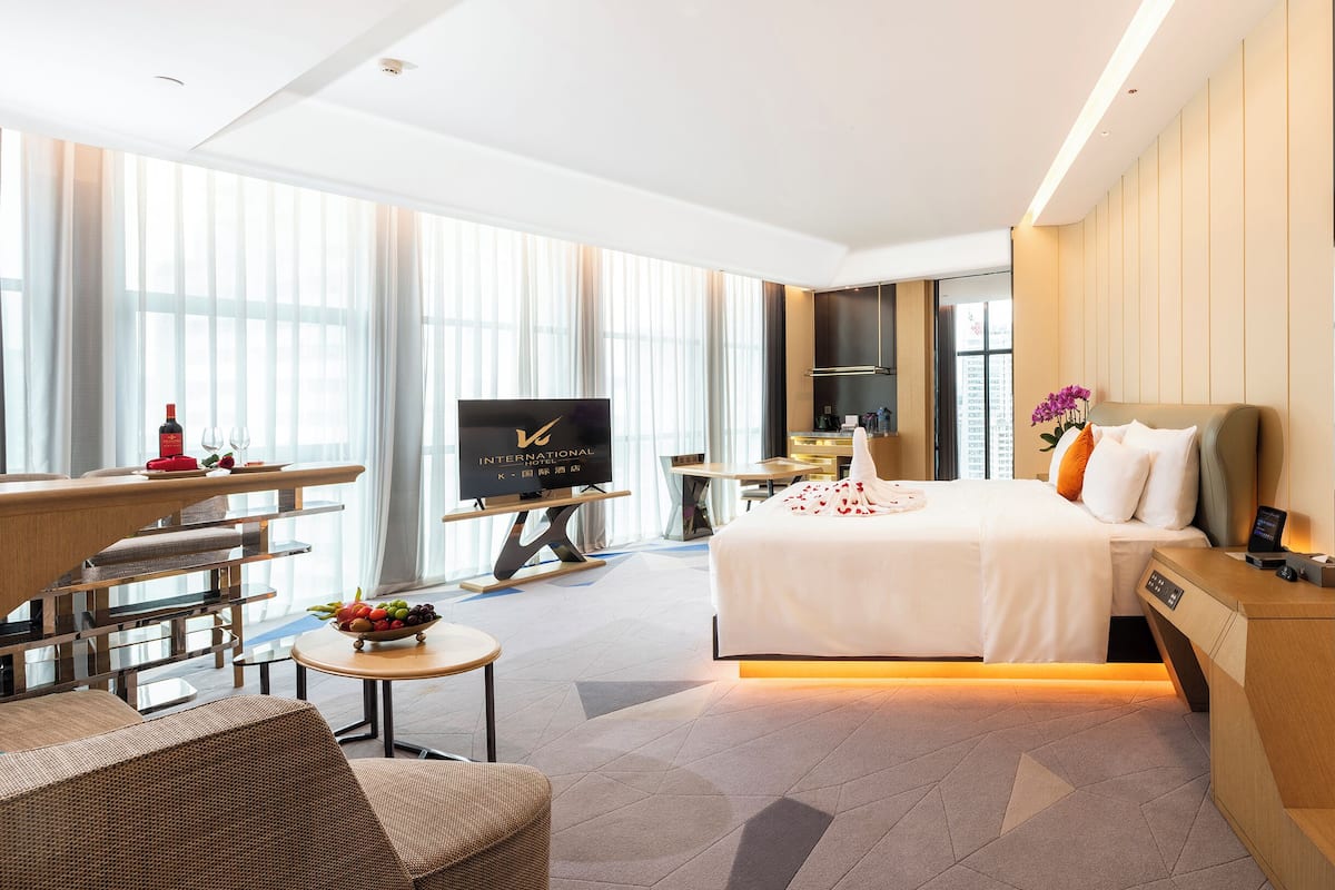 A modern hotel room with a large bed, a desk with a flat-screen TV, a small round table with fruit, and large windows with sheer curtains.