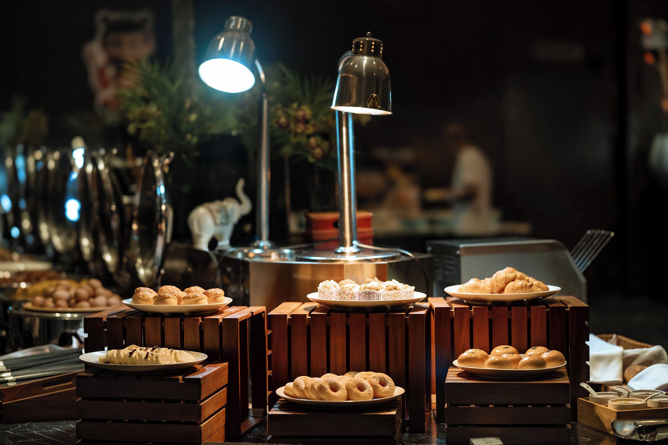 A buffet setup with various pastries and desserts displayed on wooden stands, illuminated by overhead lighting.