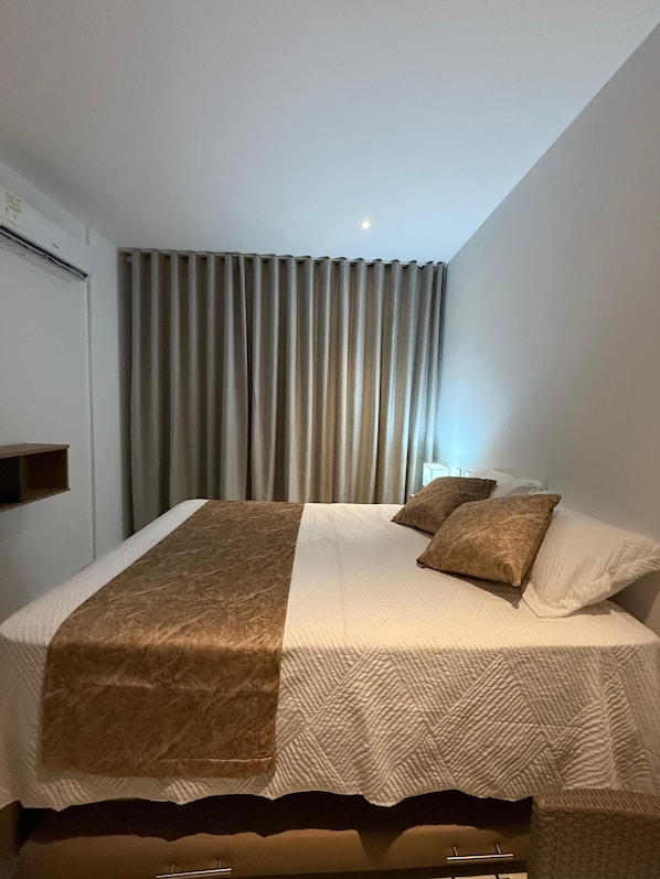 A hotel room with a bed, curtains, a bedside table, and a wall-mounted shelf.