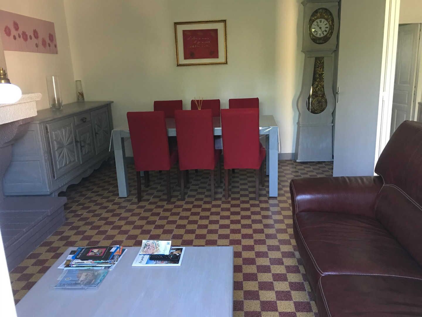 Un salon avec un sol à carreaux, un canapé en cuir marron, une table à manger avec des chaises rouges, une horloge et un tableau encadré accroché au mur.