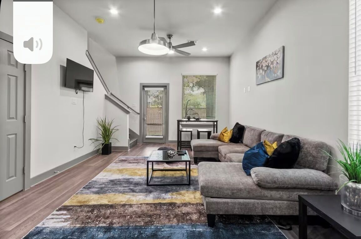 Un salon moderne avec un canapé gris, une table basse, un téléviseur fixé au mur, un ventilateur de plafond et un escalier menant à un autre niveau.
