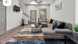 A modern living room with a grey sofa, a coffee table, a TV mounted on the wall, a ceiling fan, and a staircase leading to another level. - Modern 3BR Home w/Private Yard + Garage (Houston)