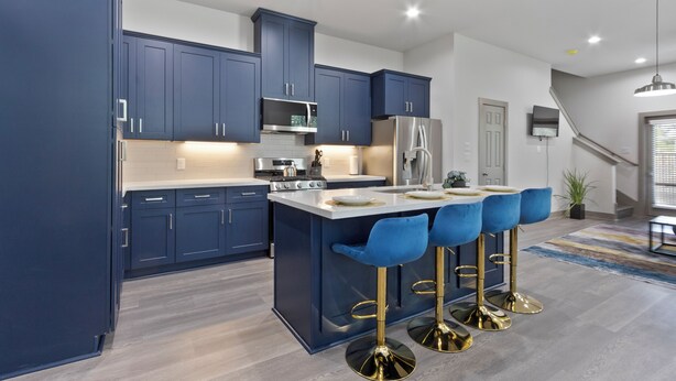 A modern kitchen with dark blue cabinets, a central island with bar stools, stainless steel appliances, and a staircase leading to another level. - Modern 3BR Home w/Private Yard + Garage (Houston)