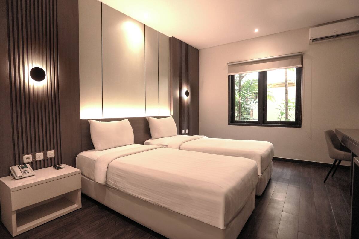 Une chambre d’hôtel moderne avec deux lits, une table de chevet avec un téléphone et une fenêtre donnant sur des palmiers.