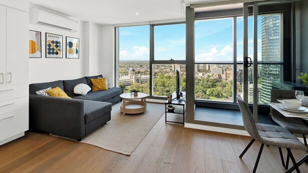 A modern living room with a sofa, coffee table, and dining area with a view of a cityscape. - Marvellous Views - Bright & Spacious CBD Escape (Melbourne)