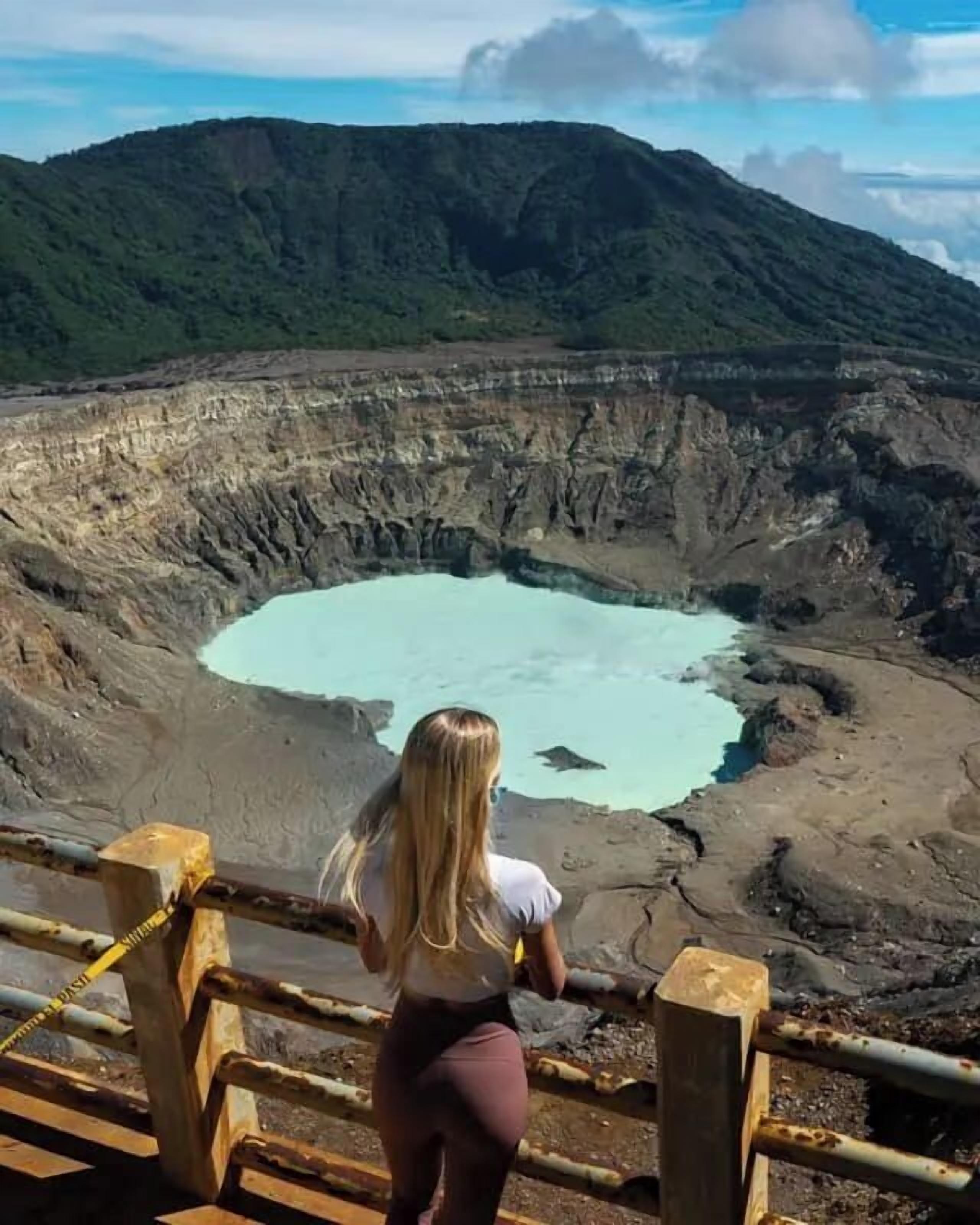 Une personne se tient sur une plateforme d’observation, d’où l’on peut admirer un vaste cratère et un lac aux eaux turquoise.