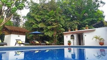 A swimming pool with lounge chairs, a small building with a door, and lush greenery in the background.
