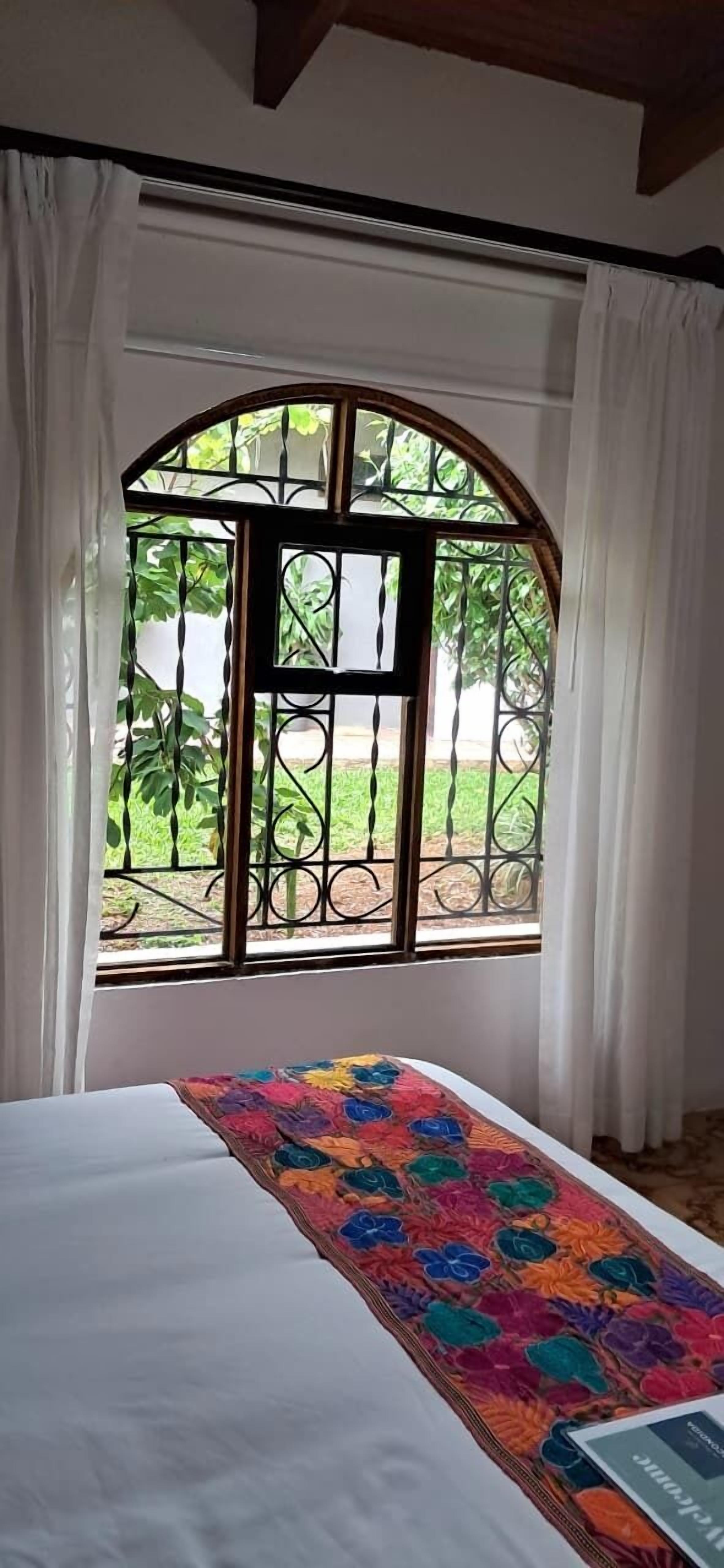 A room with a bed, a window with a metal gate, and a view of greenery outside.