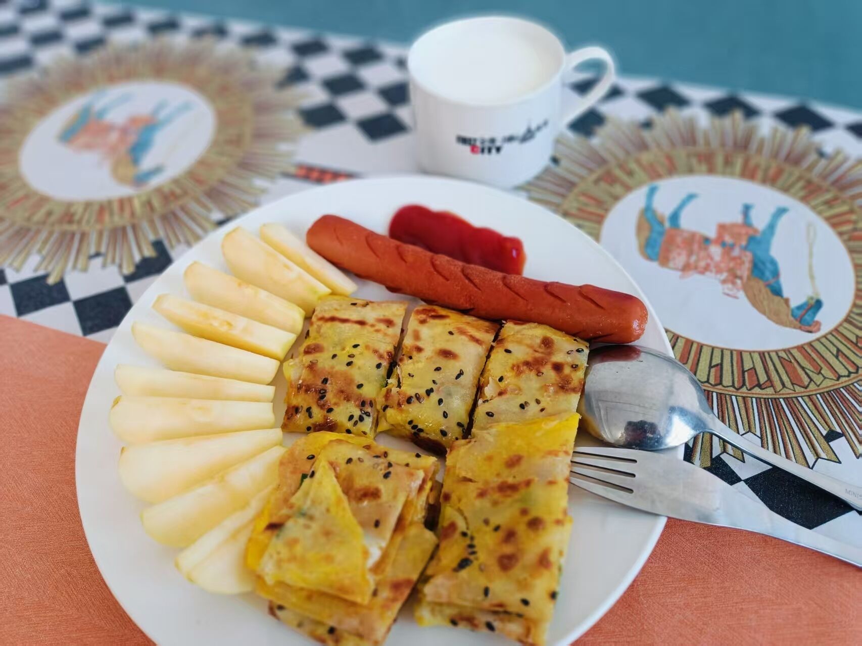 Ein Teller mit Scheibenpotatoes, eine gegrillte Pattie und ein Hotdog, begleitet von einer weißen Tasse und einem Löffel auf einer karierten Tischdecke.