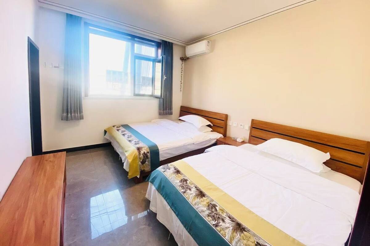A hotel room with two single beds, a wooden desk, a window with curtains, and an air conditioning unit.