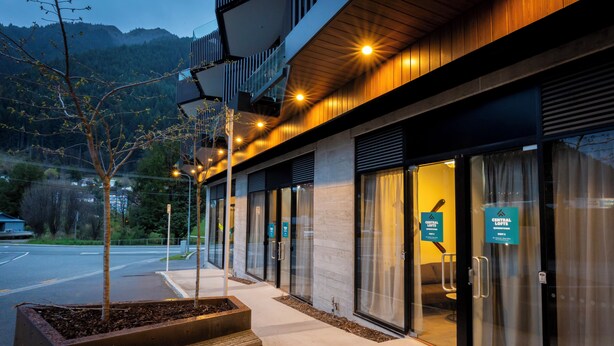 A modern hotel entrance with glass doors, a wooden bench, and a landscaped area with trees. - Central Lofts 2 & 3 - Brand New! (Queenstown)