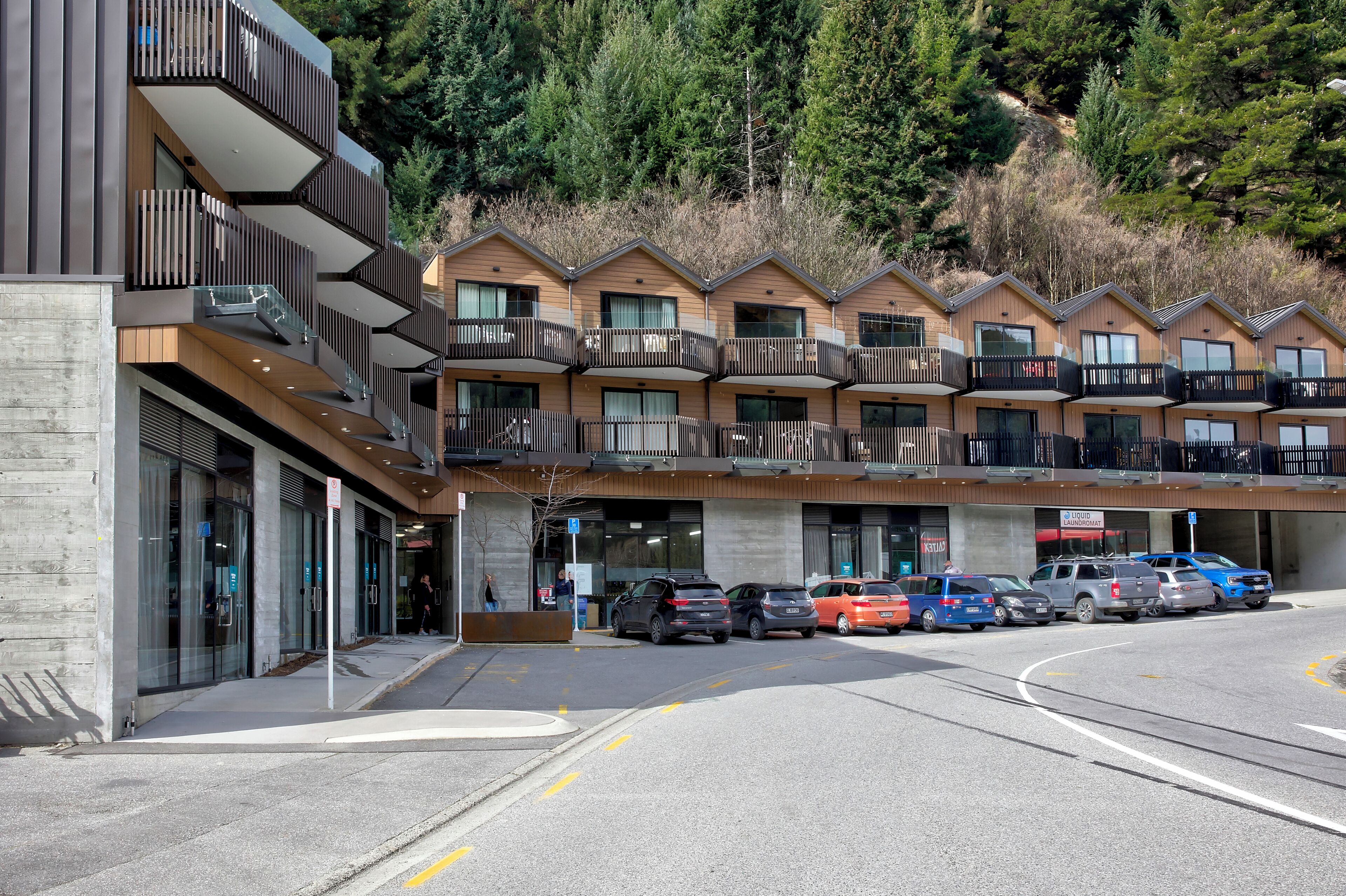 A modern hotel with multiple balconies, a parking lot, and a view of a forested hillside.