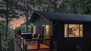 Una cabaña con terraza, una cocina visible a través del ventanal y un paisaje boscoso de fondo.