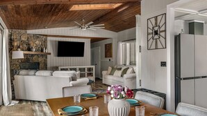 Una sala de estar moderna con techo de madera, paredes blancas, una pared de acento de piedra, un televisor de pantalla plana, una zona de comedor con una mesa de madera para cuatro, y un área de cocina con refrigerador.