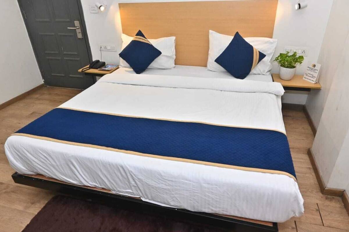 Un lit bien fait, recouvert d’une couvre-lit blanche et bleue, avec une tête de lit en bois et une table de chevet agrémentée d’une plante et d’un petit panneau.