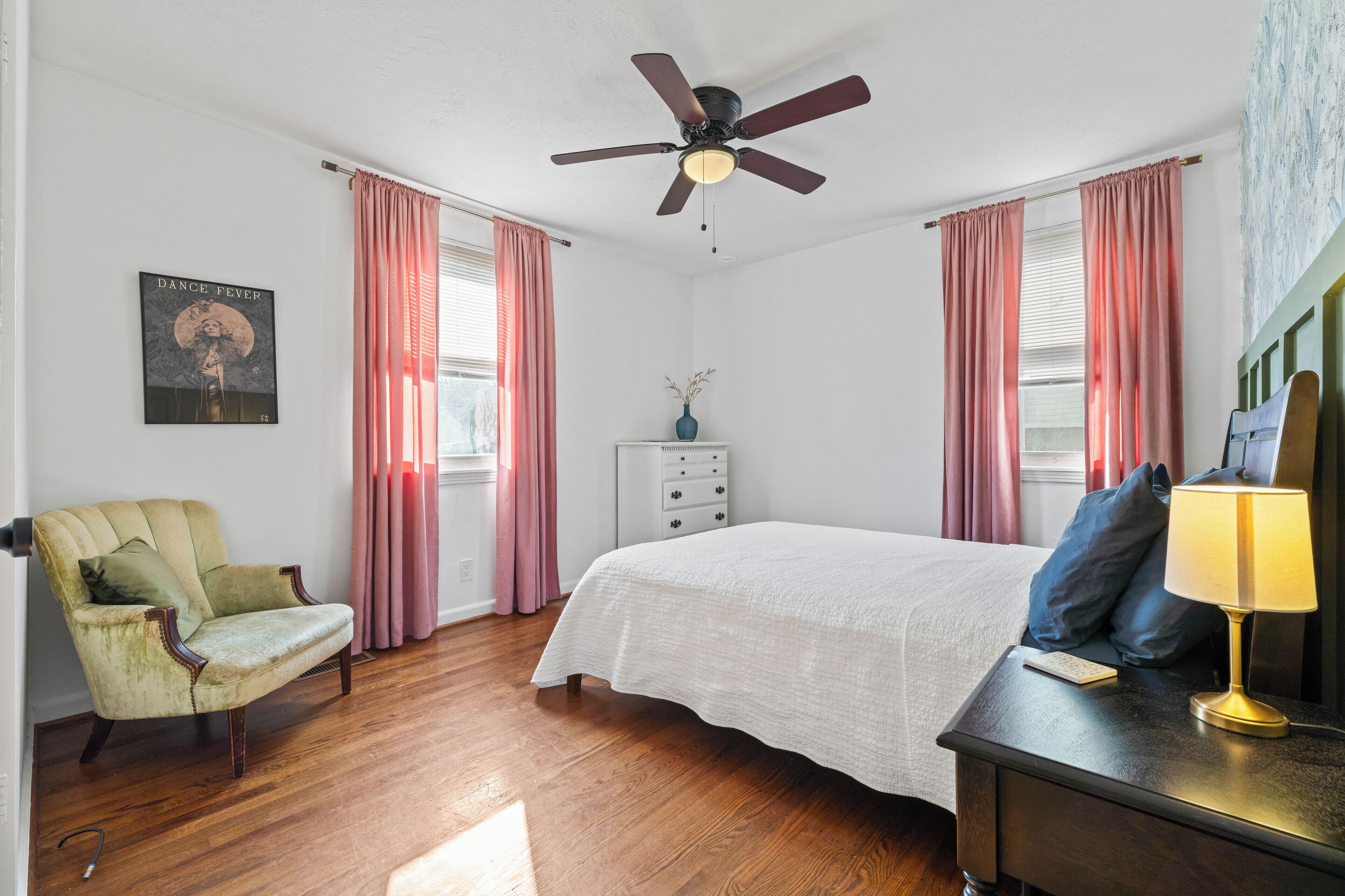 A bedroom with a bed, a chair, a dresser, a ceiling fan, and a window with curtains.