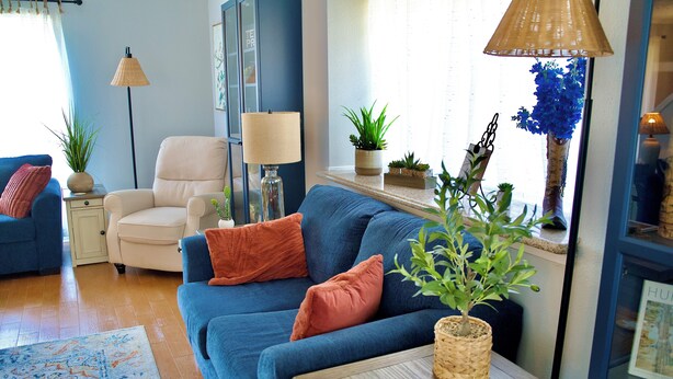 A living room with a blue sofa, a white armchair, and a small wooden table with a plant. There is a floor lamp and a window with blue curtains. - The Elisha House - Cozy Stay Near Local Eats (Katy)