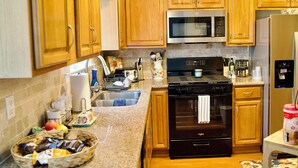 A kitchen with wooden cabinets, a stainless steel microwave, a black stove, and a refrigerator. - The Elisha House - Cozy Stay Near Local Eats (Katy)