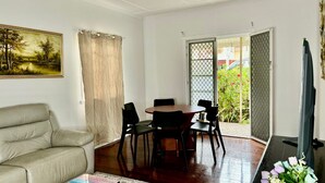 A living room with a sofa, a dining table with chairs, a TV, and a view of greenery outside. - Affordable Chermside Home near Hospital & Westfield (Chermside)