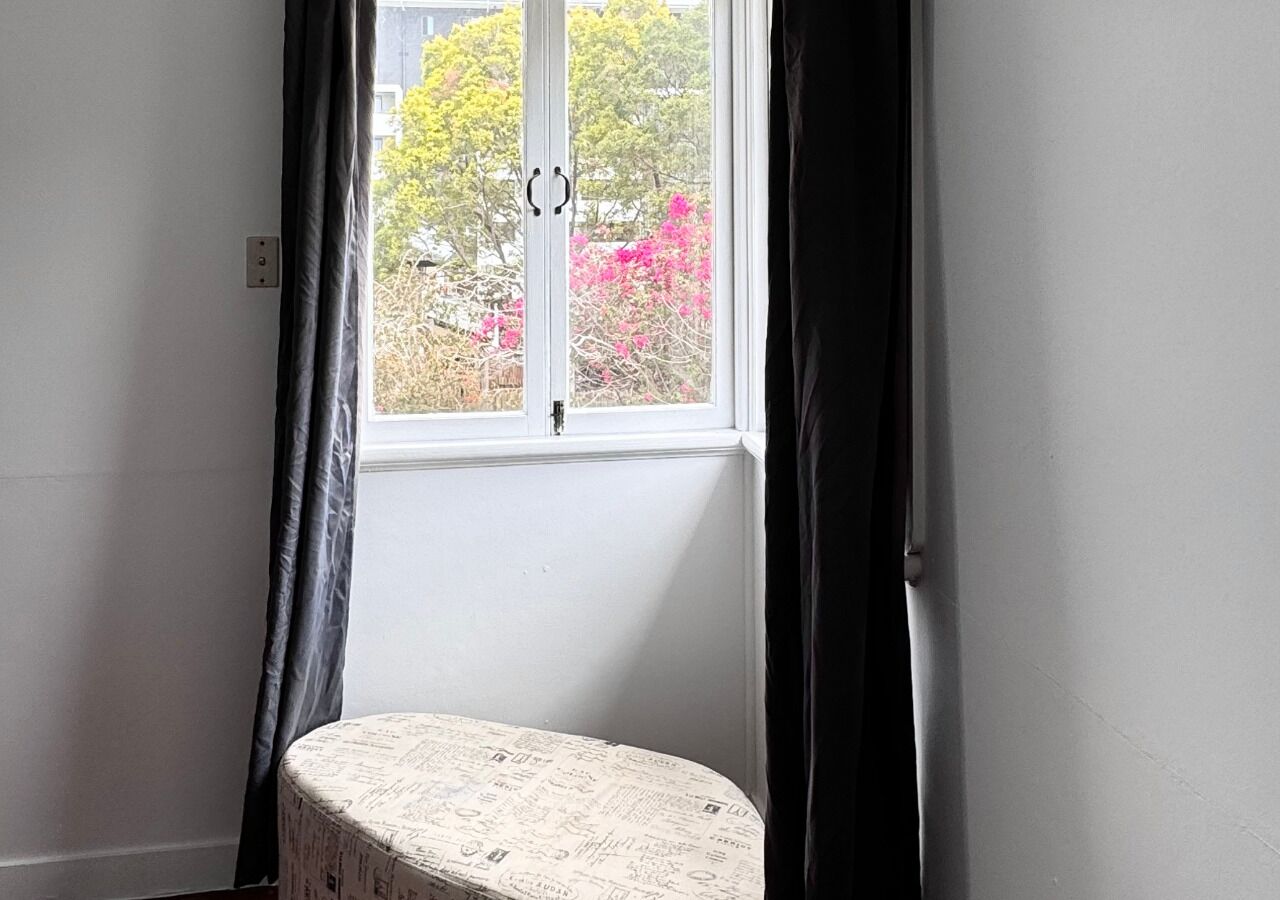 A corner room with a small round table, a window with curtains, and a view of trees and buildings outside.