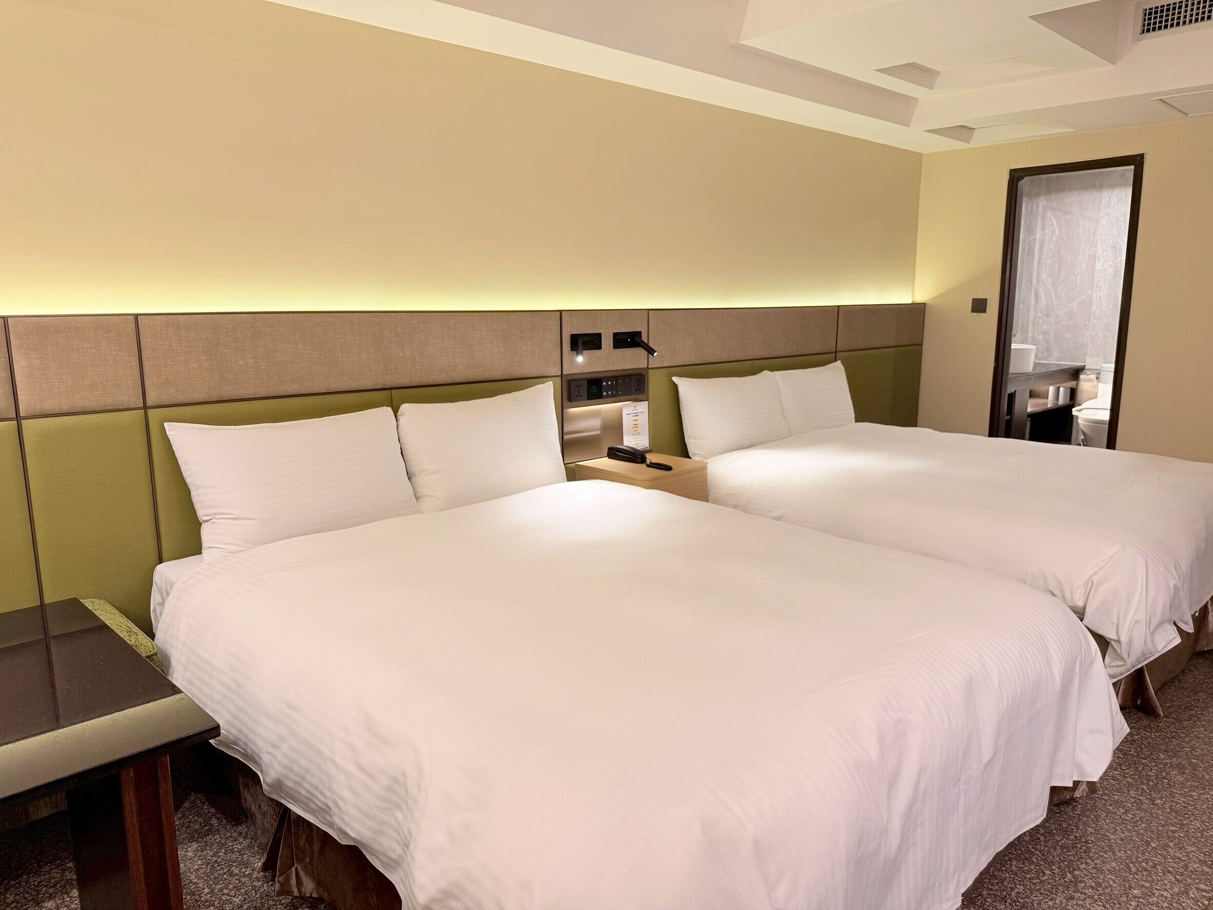 A hotel room with two beds, a bedside table, a mirror, and a wall-mounted control panel.