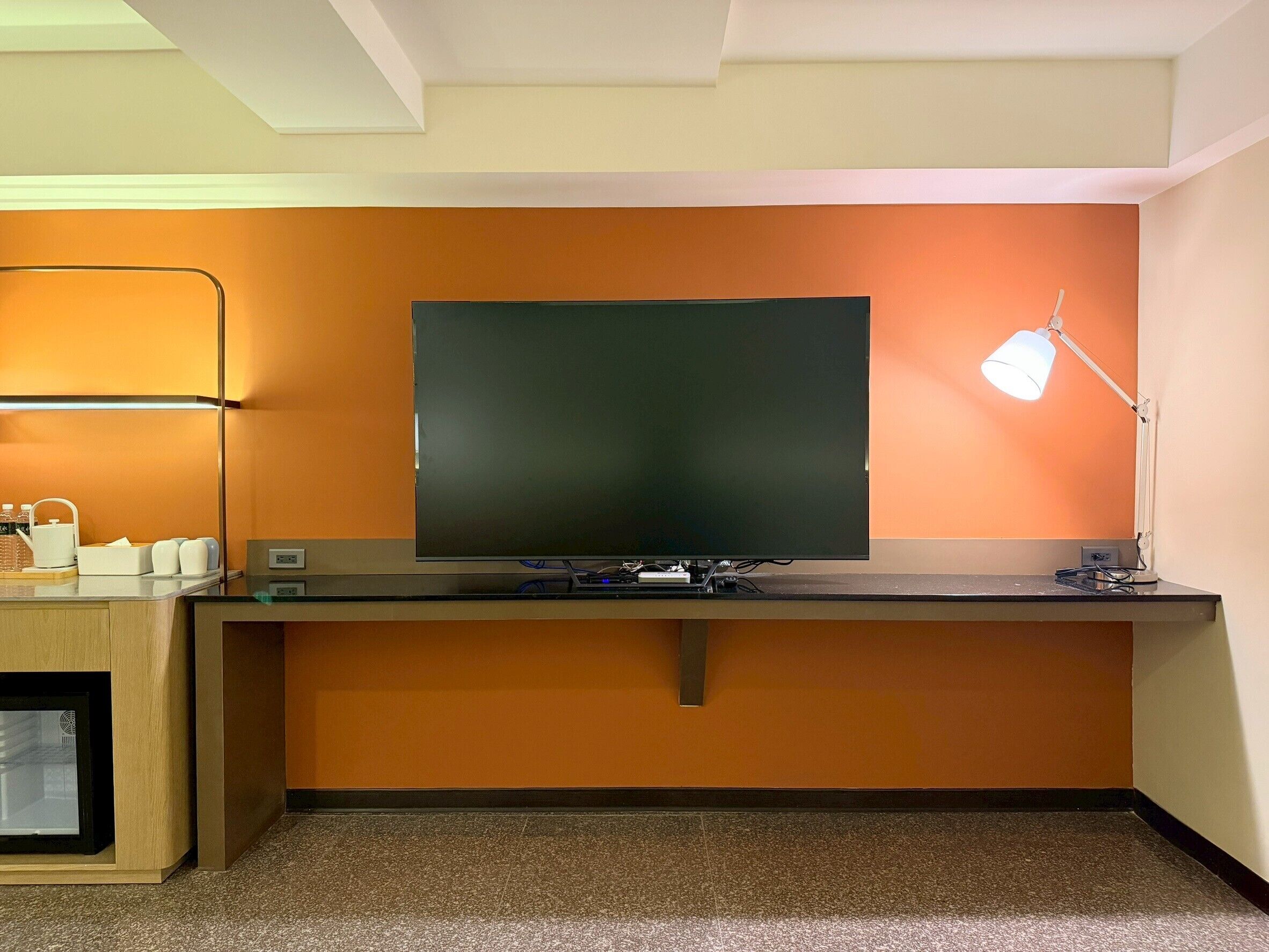 A room with a flat-screen TV mounted on a wall, a desk with a lamp, and a small refrigerator.