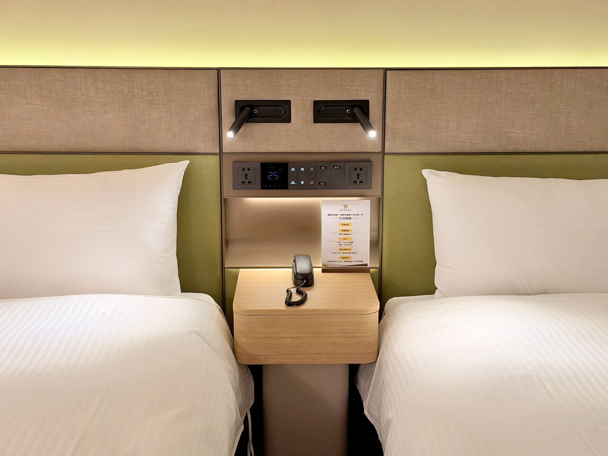 A hotel room with two beds, a bedside table with a phone, a digital clock, and a sign with room information.