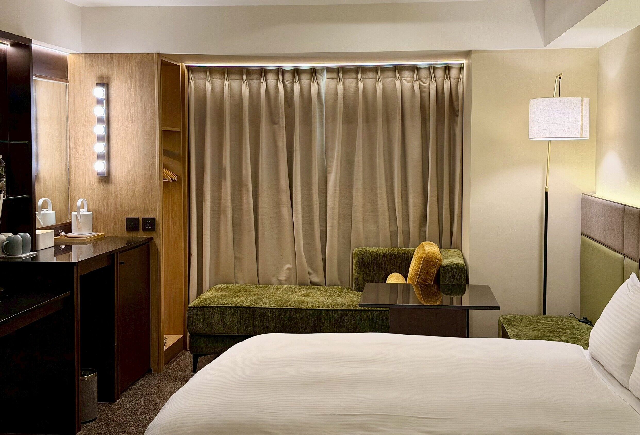 A hotel room with a bed, a desk with a mirror, a chair, a lamp, and a window with curtains.