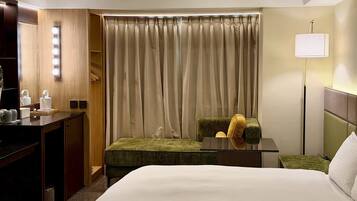 A hotel room with a bed, a desk with a mirror, a chair, a lamp, and a window with curtains.