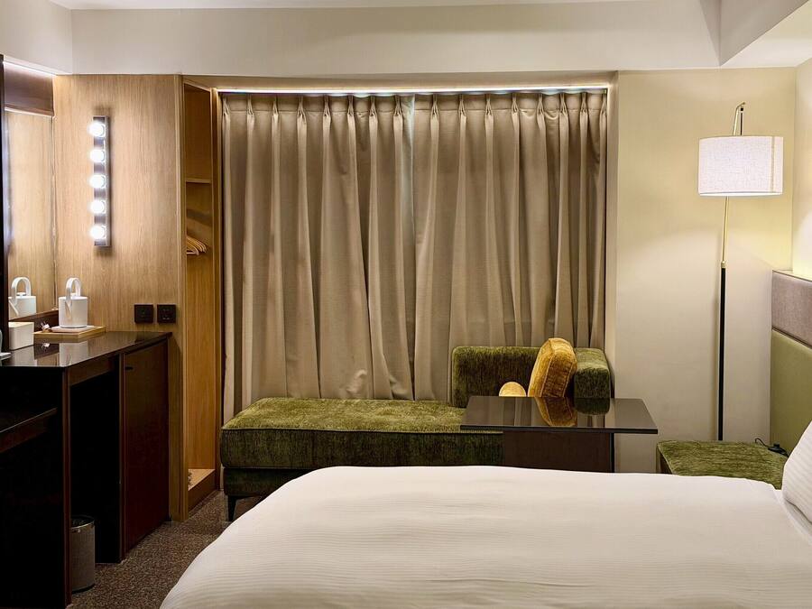 A hotel room with a bed, a desk with a mirror, a chair, a lamp, and a window with curtains.