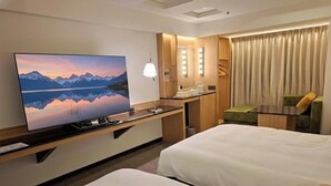 Habitación de hotel con un televisor de pantalla plana grande, dos camas, un escritorio con espejo y vistas a montañas reflejadas en la ventana.