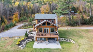 A two-story log cabin with a wrap-around porch, stone foundation, and a view of a forested area. - Beautiful Custom Log Home - 1 Mile to Sunday River, Walk to Matterhorn (Newry)