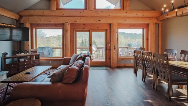 A cozy cabin interior with a wooden dining table, a sofa, and a TV. Large windows offer views of the outdoors. - Beautiful Custom Log Home - 1 Mile to Sunday River, Walk to Matterhorn (Newry)